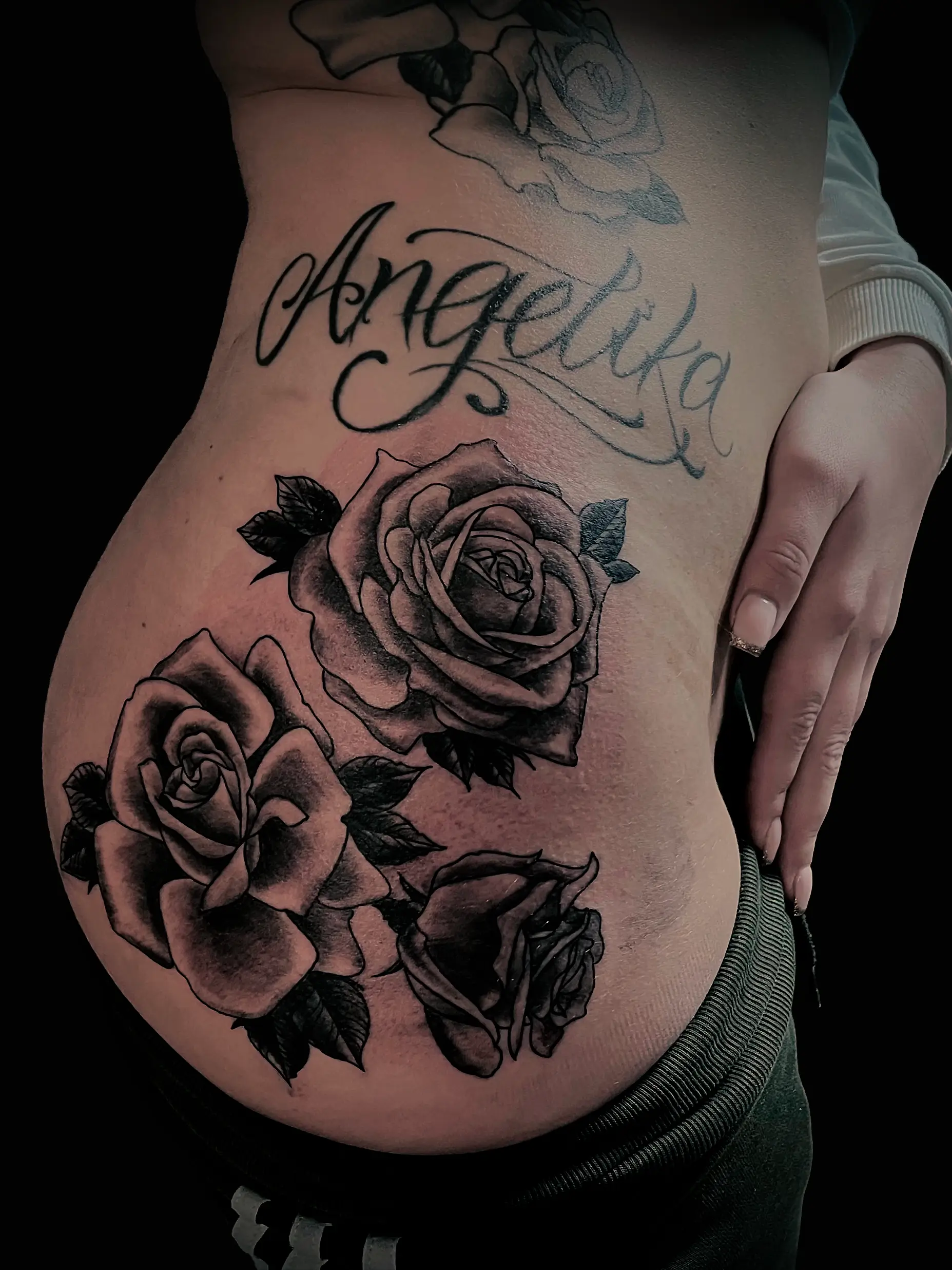 Script tattoo “Angelika” combined with realistic black and grey roses on hip.