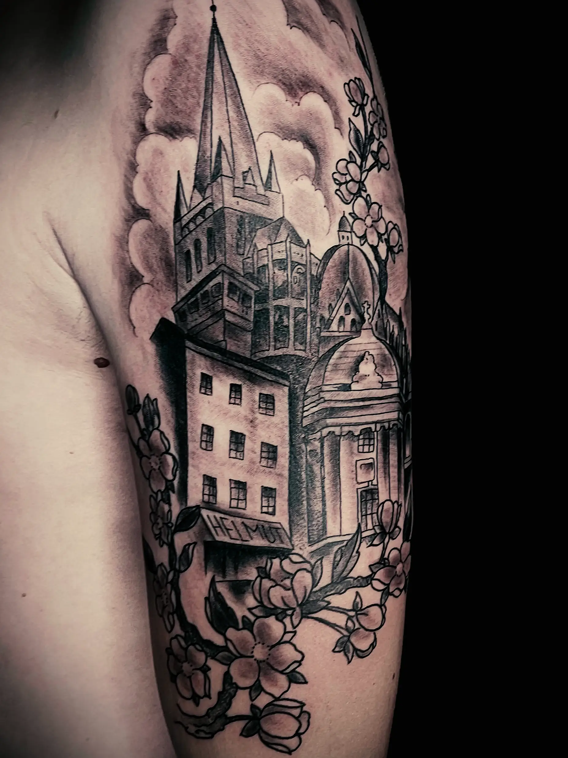 Black and grey tattoo of architectural buildings with floral elements, detailed shading on upper arm.
