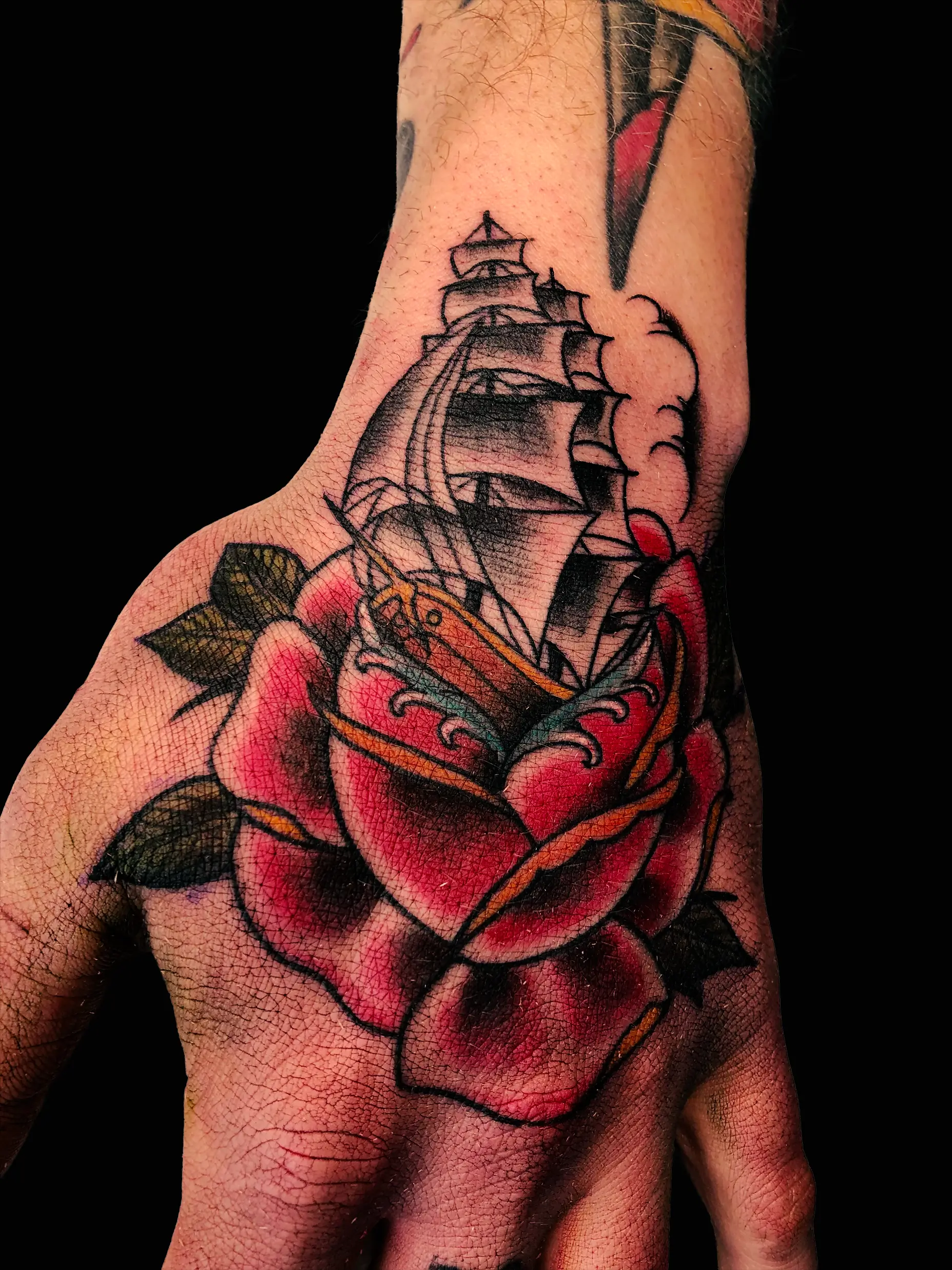 Traditional style tattoo of a sailing ship emerging from a red rose on the back of a hand, bold outlines and color accents.