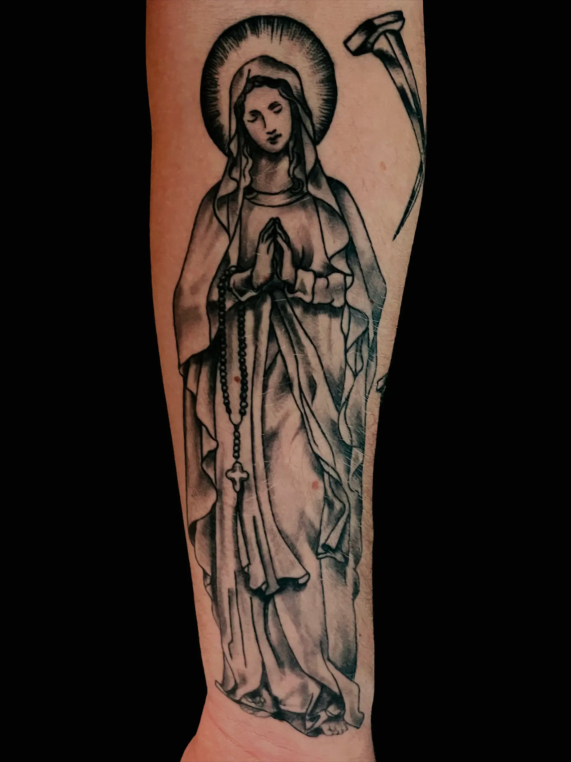 Black and grey tattoo of a praying Madonna with rosary and halo, placed on lower leg.