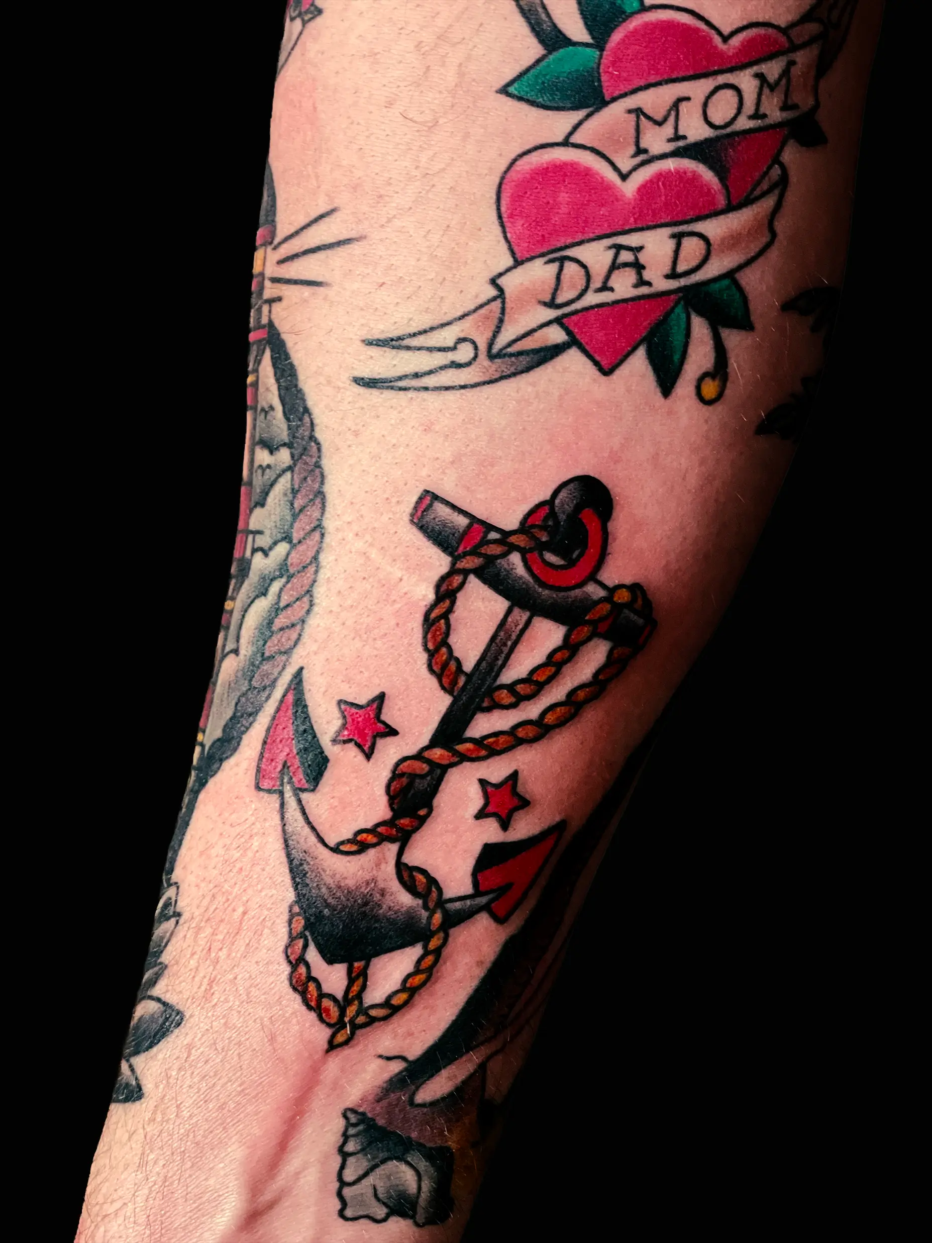 Traditional tattoo with red hearts and banner reading “Mom Dad”, combined with anchor and rope elements on arm.