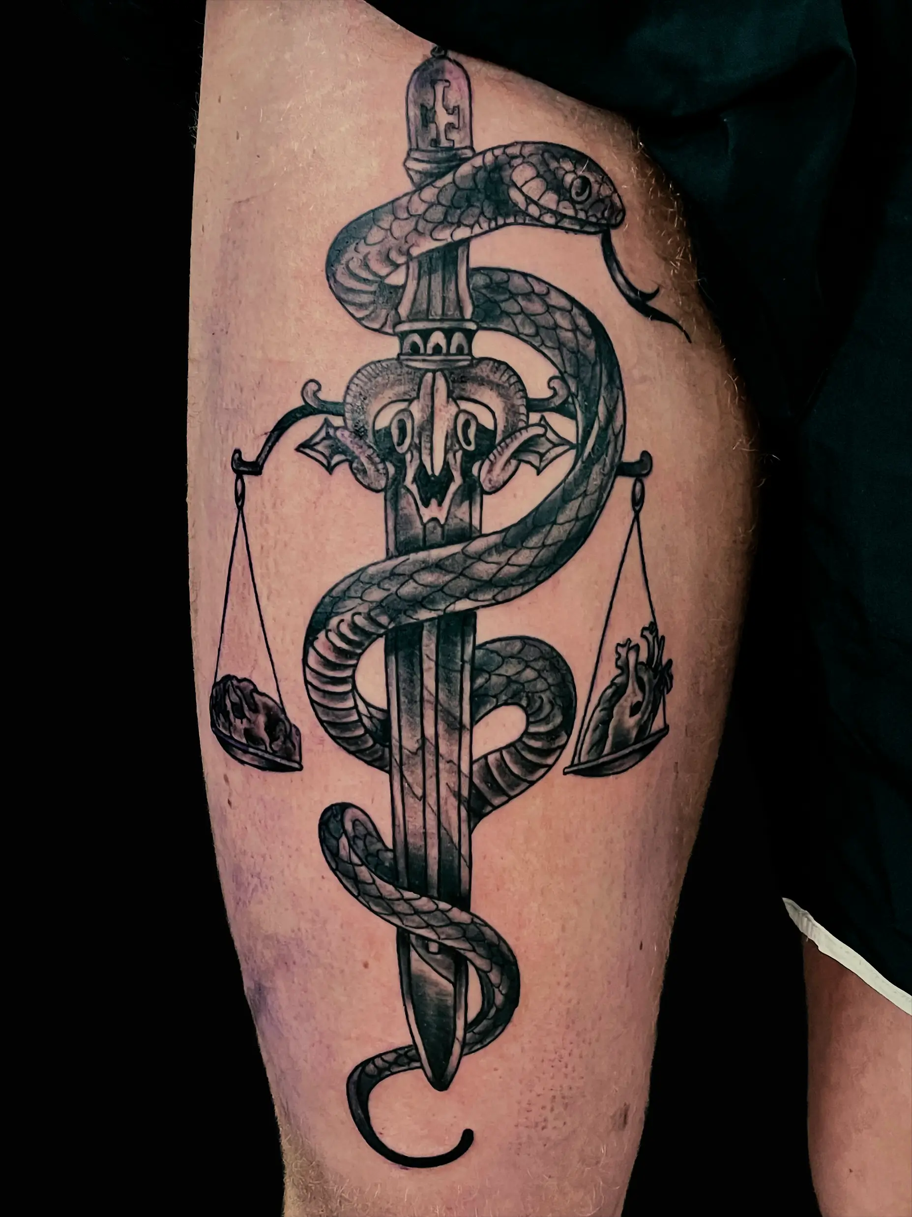 Black and grey tattoo of a snake wrapped around a dagger with scales of justice, placed on thigh.